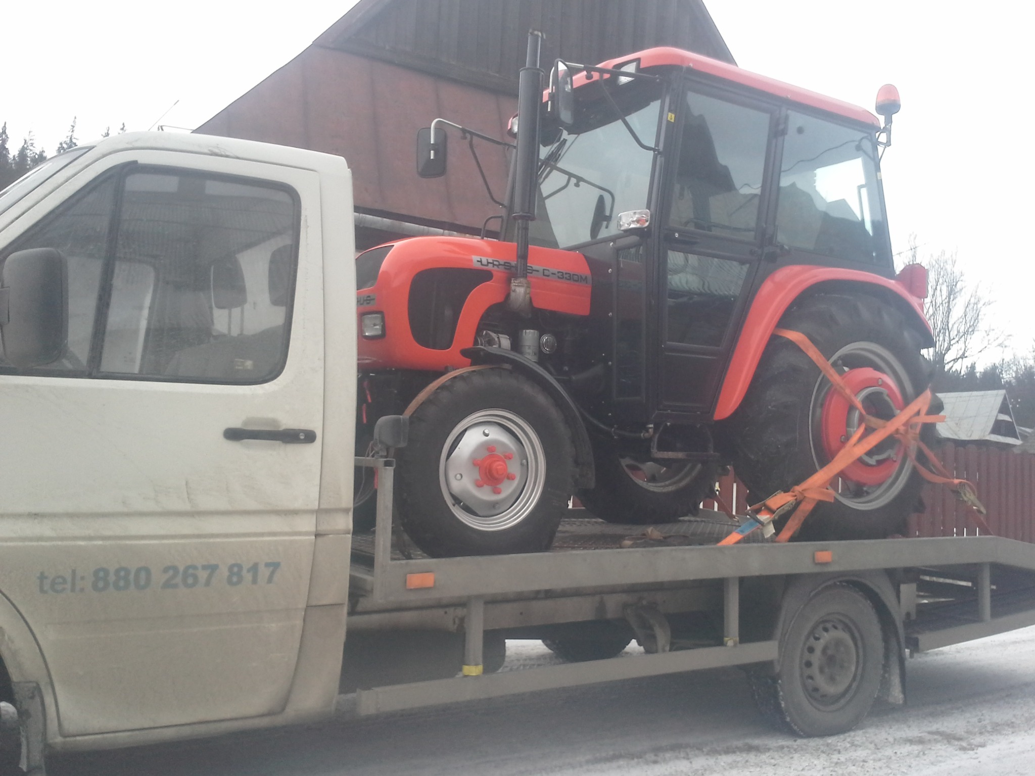 Czerwony traktor Ursus C-330M zabezpieczony pomarańczowymi pasami na platformie lawety, z widocznym numerem telefonu na brudnym białym samochodzie holowniczym i fragmentem budynku w tle.