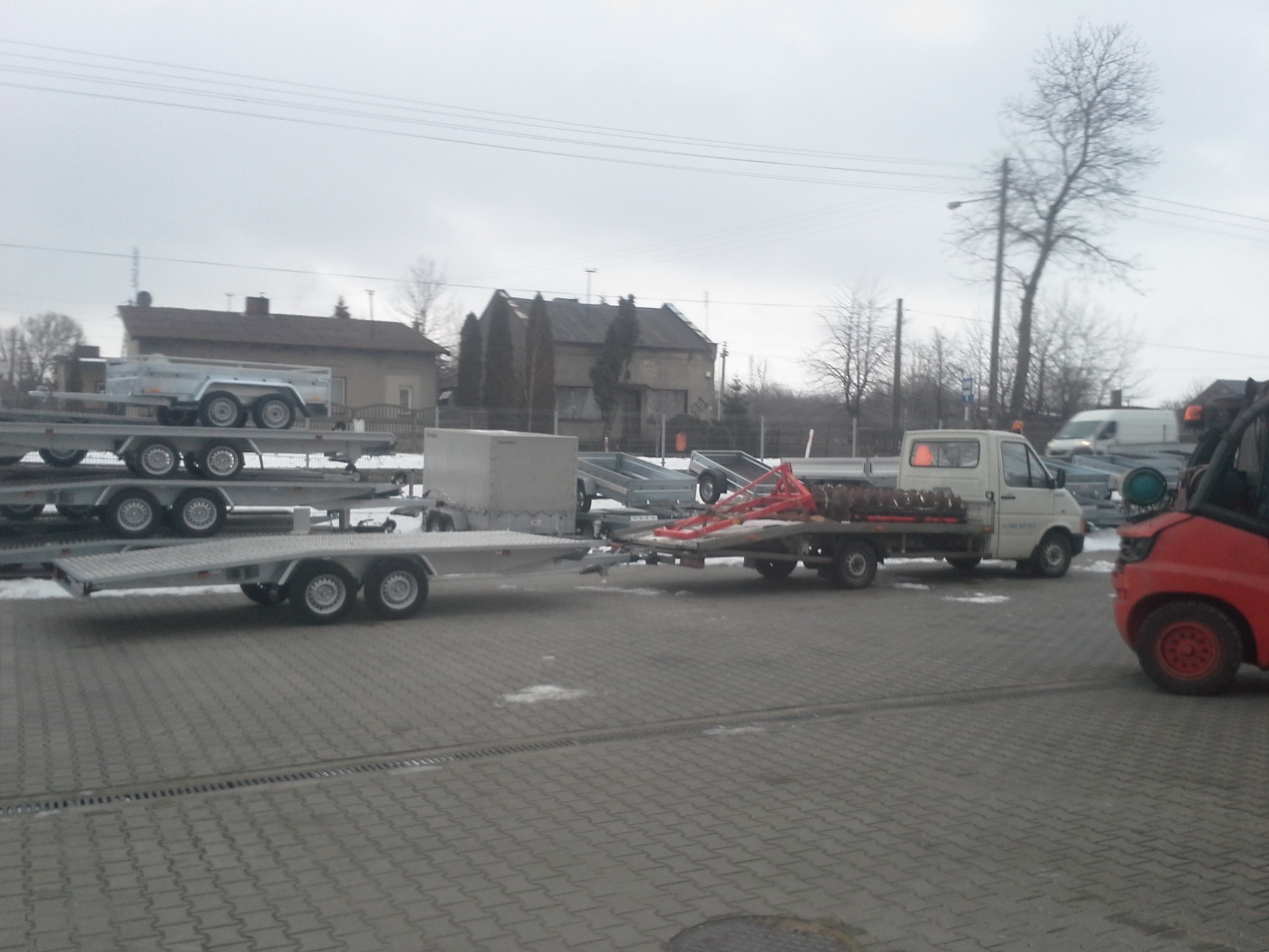 Prezentacja nowych przyczep samochodowych na placu, widoczny biały samochód dostawczy z przyczepą oraz czerwony wózek widłowy.