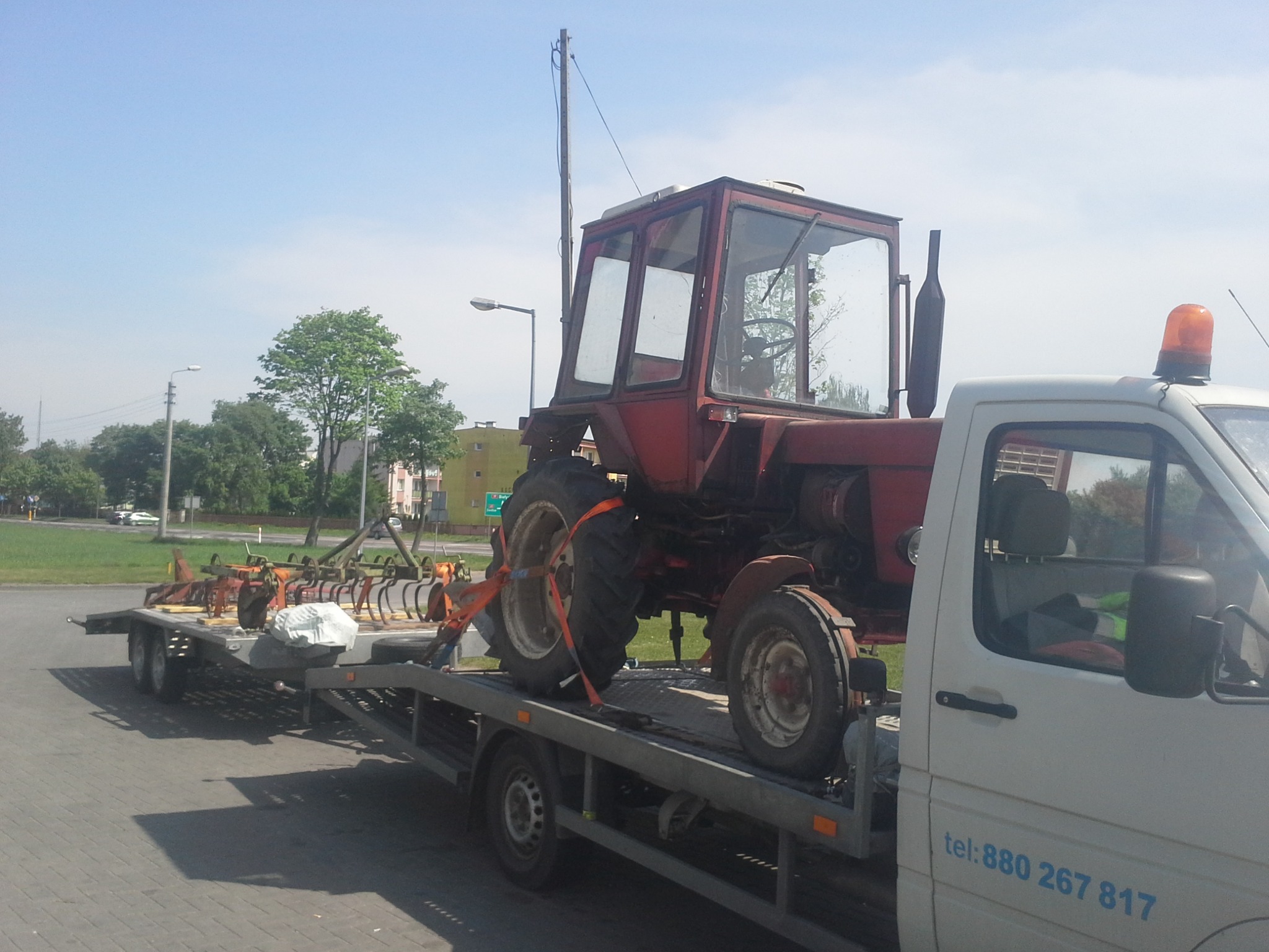 Czerwony traktor z kabiną zabezpieczony pasami na lawecie z dodatkowym sprzętem rolniczym, widok z boku na tle miejskiego krajobrazu.