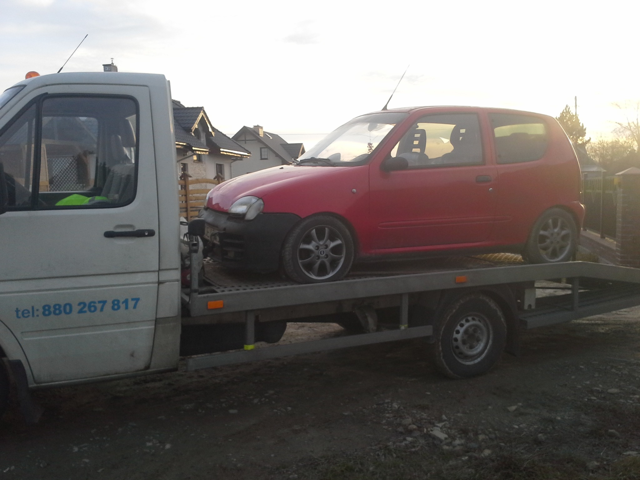 Biała laweta z czerwonym samochodem osobowym na platformie, zaparkowana na grząskim terenie, w tle domy jednorodzinne.