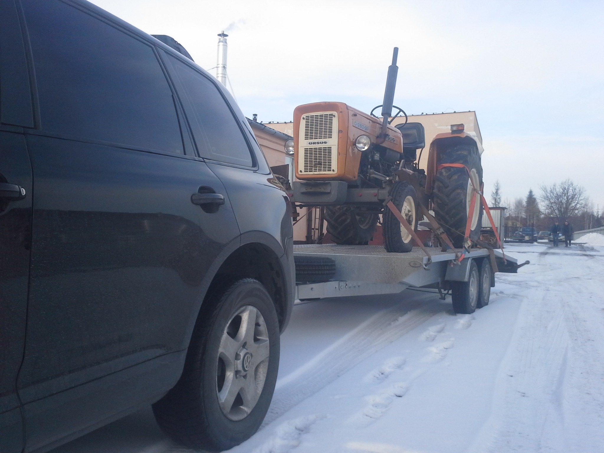 Czarny SUV holuje na przyczepie traktor Ursus w zimowej scenerii, widoczne ślady opon na śniegu.