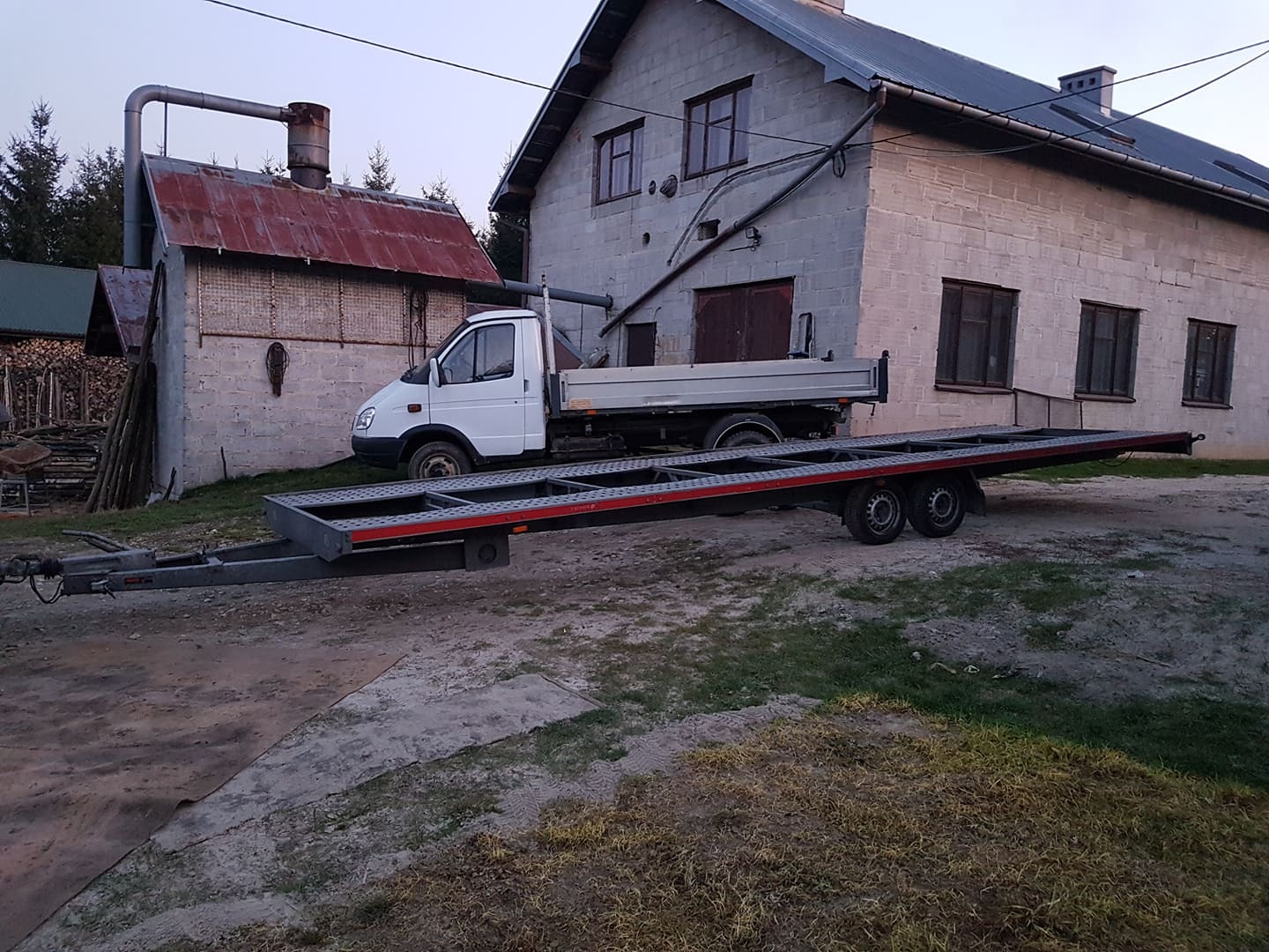 Biały samochód dostawczy z przyczepą lawetą na posesji w Szerzynach, z budynkami w tle.