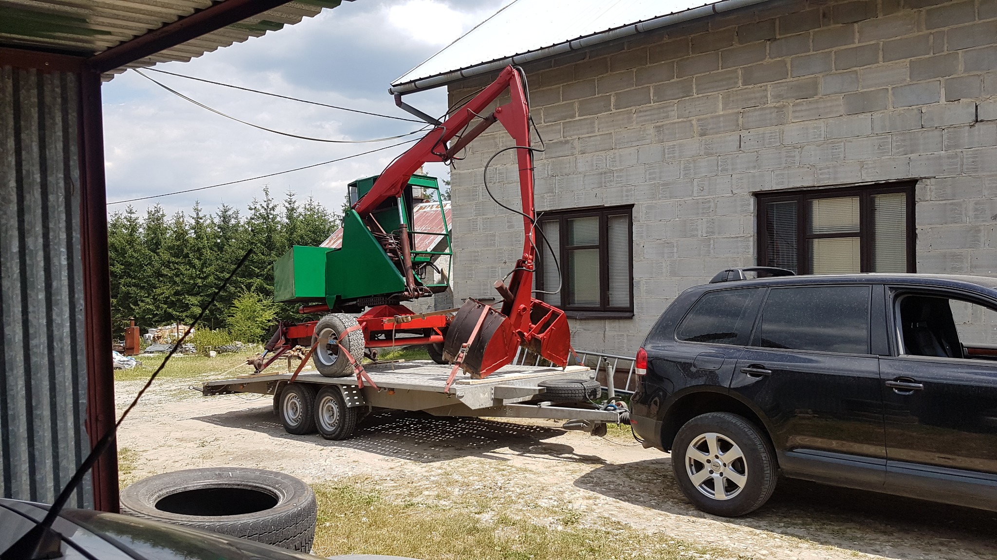 Czerwono-zielona maszyna z łyżką na przyczepie, ciągnięta przez czarny samochód terenowy na tle murowanego budynku z oknami i lasu iglastego.