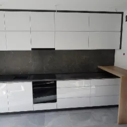 Biała kuchnia z połyskiem, szare marmurowe płytki nad blatem, drewniany barek, szare płytki podłogowe, czarne obramowanie szafek górnych.