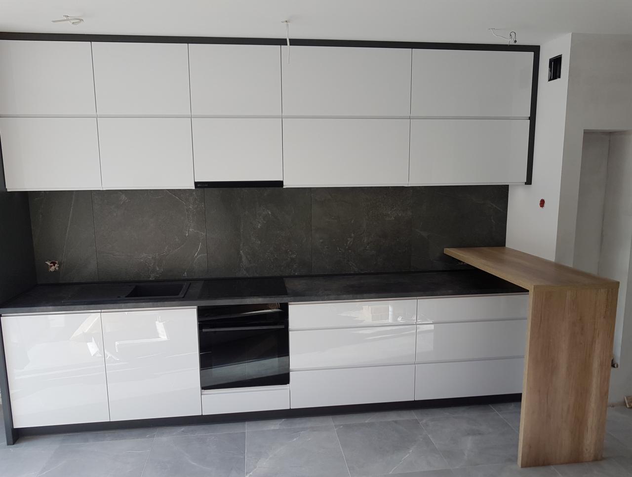 Biała kuchnia z połyskiem, szare marmurowe płytki nad blatem, drewniany barek, szare płytki podłogowe, czarne obramowanie szafek górnych.
