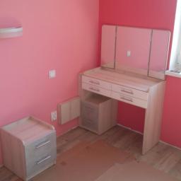 Js Kama - Jasna toaletka z trzema lustrami i szafka nocna na tle różowych ścian, zabezpieczone kartonem podłoże, w trakcie montażu.