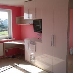 Js Kama - Jasna zabudowa meblowa z szafą, biurkiem i półkami w pokoju z różowymi ścianami, widoczne zabezpieczenie podłogi kartonem.