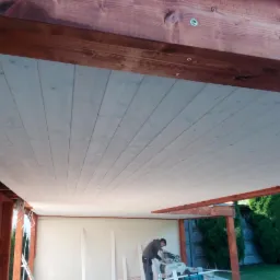 Montaż drewnianego zadaszenia tarasu: widok od dołu na deski pokryte jasną bejcą, mocowane do brązowych belek konstrukcyjnych; w tle pracownik przycina deskę za pomocą piły stołowej.