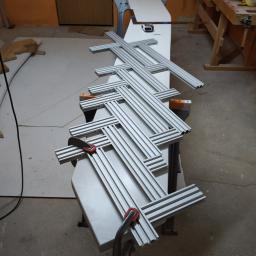 Js Kama - Elementy aluminiowe profilowane, częściowo skręcone, leżą na stole warsztatowym, przygotowane do montażu konstrukcji; w tle widoczny warsztat z narzędziami i materiałami.