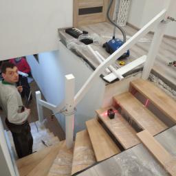 Js Kama - Montaż drewnianych schodów: widok z góry na pracownika z wiertarką, laserowy poziom na stopniach zabezpieczonych folią, biała balustrada w trakcie instalacji, narzędzia i odkurzacz w tle.