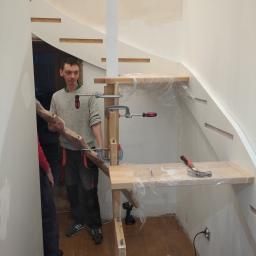 Js Kama - Stolarz trzyma element drewniany podczas montażu schodów z drewnianymi stopniami, zabezpieczonymi folią, w trakcie prac wykończeniowych.