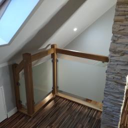 Js Kama - Szklana balustrada z drewnianymi słupkami, zabezpieczająca antresolę na poddaszu, widok z góry na drewnianą podłogę i kamienną kolumnę.