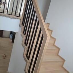 Js Kama - Jasne, drewniane schody z czarną, metalową balustradą o geometrycznym wzorze, zamontowane przy białej ścianie. Widoczne niedokończone wykończenie ściany na górze schodów.