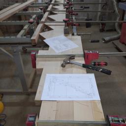 Js Kama - Deski z jasnego drewna sklejane za pomocą ścisków stolarskich, na wierzchu leżą plany techniczne i młotek.