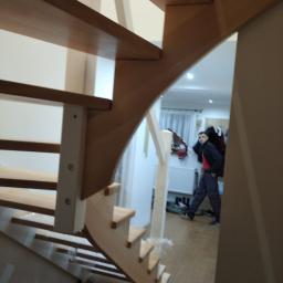 Js Kama - Drewniane schody z zaokrąglonym biegiem, widoczne z dołu, z mężczyzną w tle w pomieszczeniu z grzejnikiem i zasłonami.