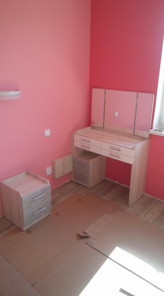 Jasna toaletka z trzema lustrami i szafka nocna na tle różowych ścian, zabezpieczone kartonem podłoże, w trakcie montażu.