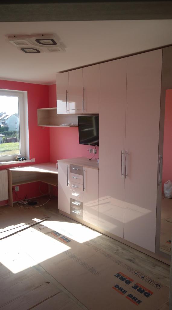 Jasna zabudowa meblowa z szafą, biurkiem i półkami w pokoju z różowymi ścianami, widoczne zabezpieczenie podłogi kartonem.