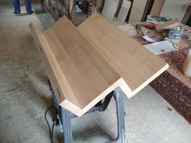 Fragment drewnianych schodów w trakcie montażu, widoczne połączenia kołkowe, na tle warsztatu z dywanem i narzędziami.