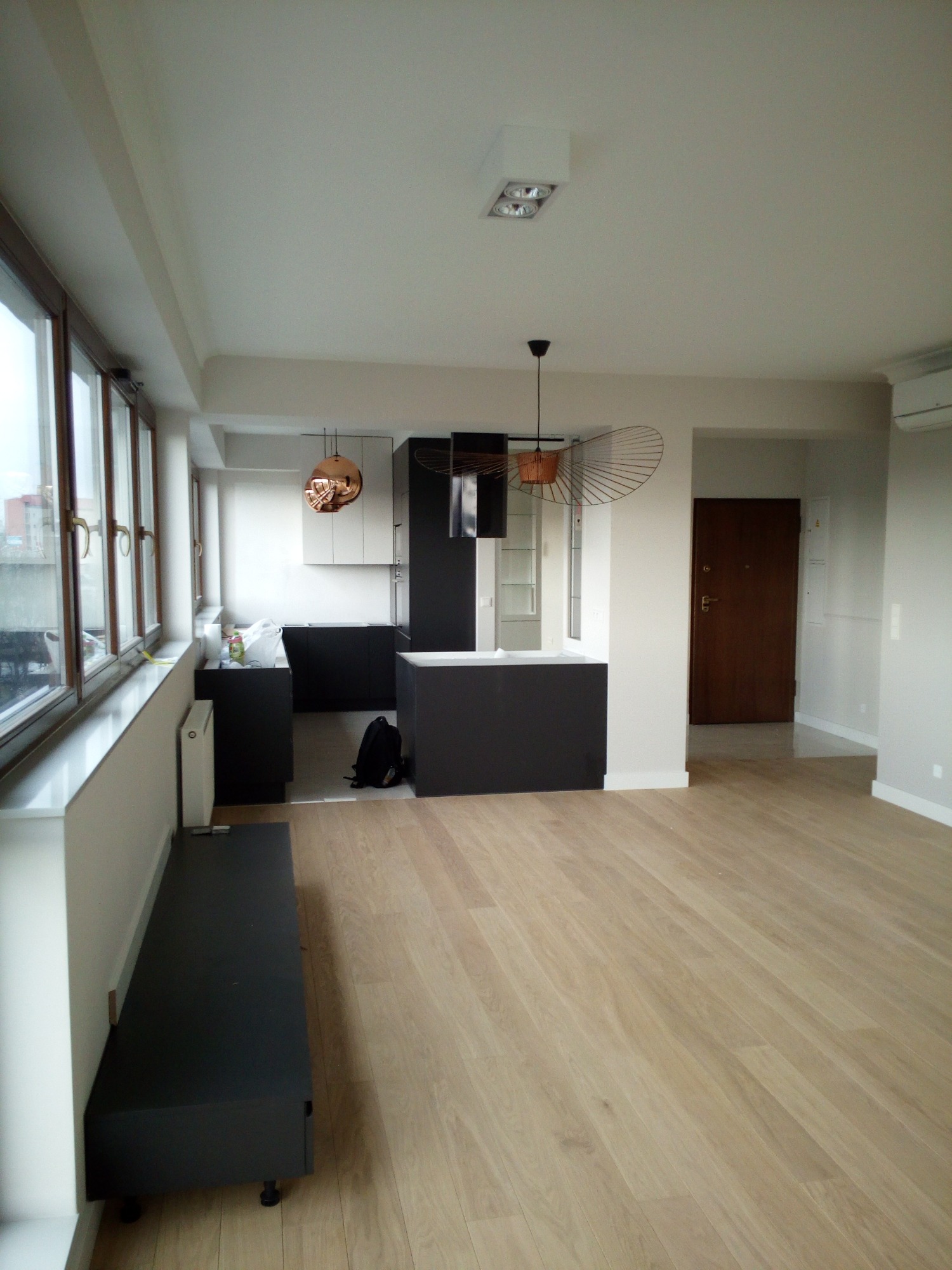 Jasne panele podłogowe w nowocześnie wykończonym wnętrzu z aneksem kuchennym, widok z perspektywy wejścia, z elementami oświetlenia dekoracyjnego i czarnymi meblami kuchennymi.