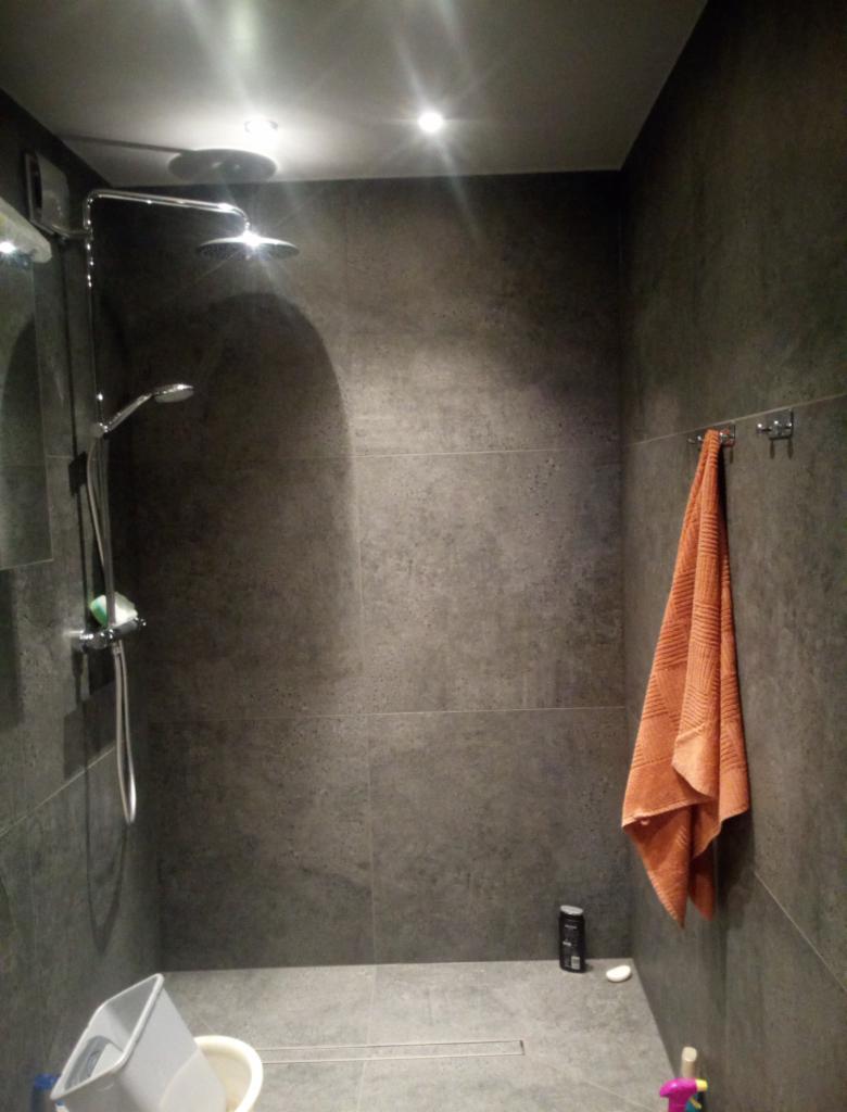 Nowoczesna kabina prysznicowa z deszczownicą i ręcznym prysznicem, wykończona szarymi płytkami imitującymi beton, z pomarańczowym ręcznikiem na wieszaku i widocznym odpływem liniowym w podłodze.