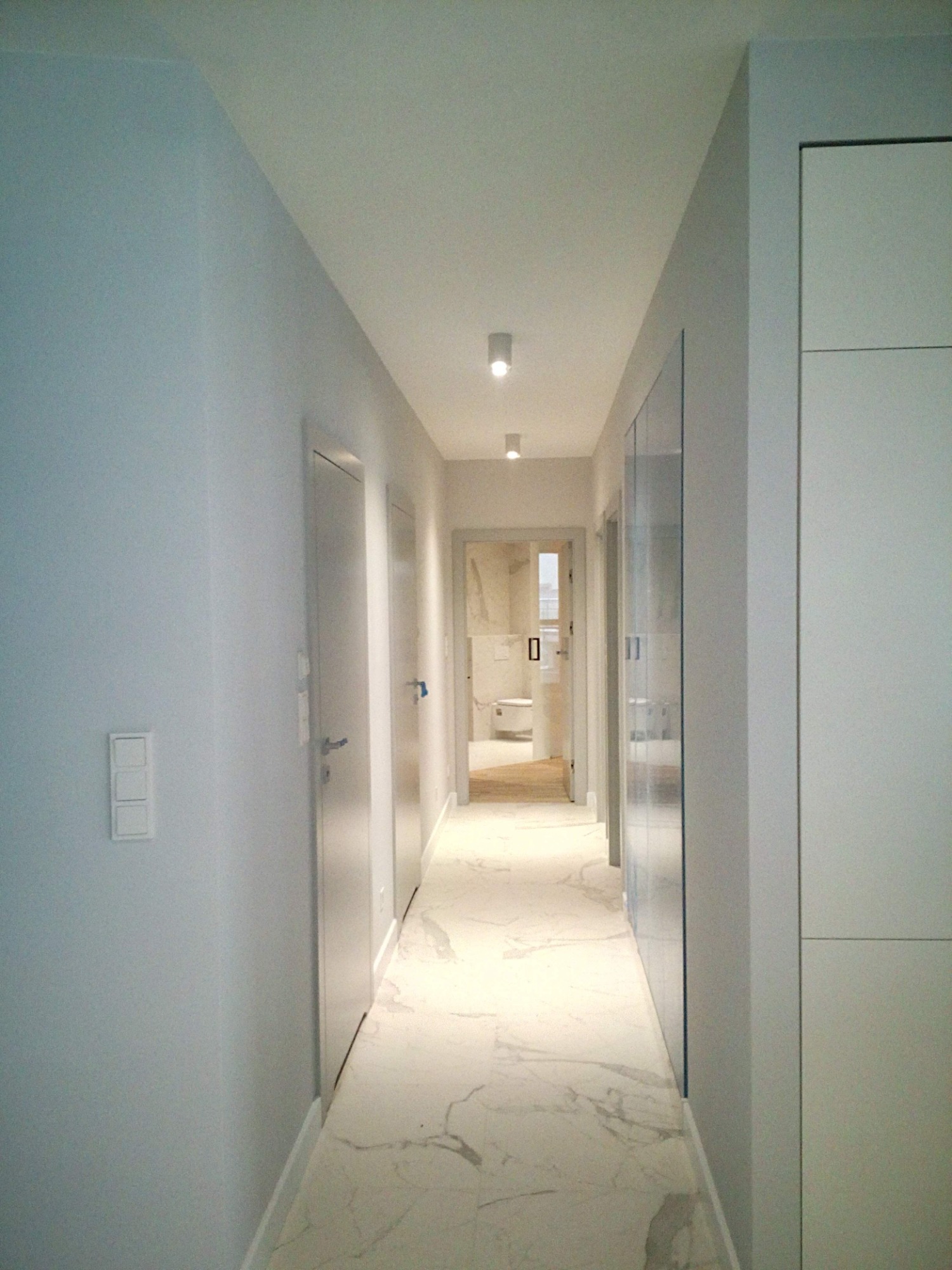 Nowoczesny korytarz z białymi ścianami, marmurową podłogą i widokiem na łazienkę w tle, oświetlony punktowymi lampami sufitowymi.