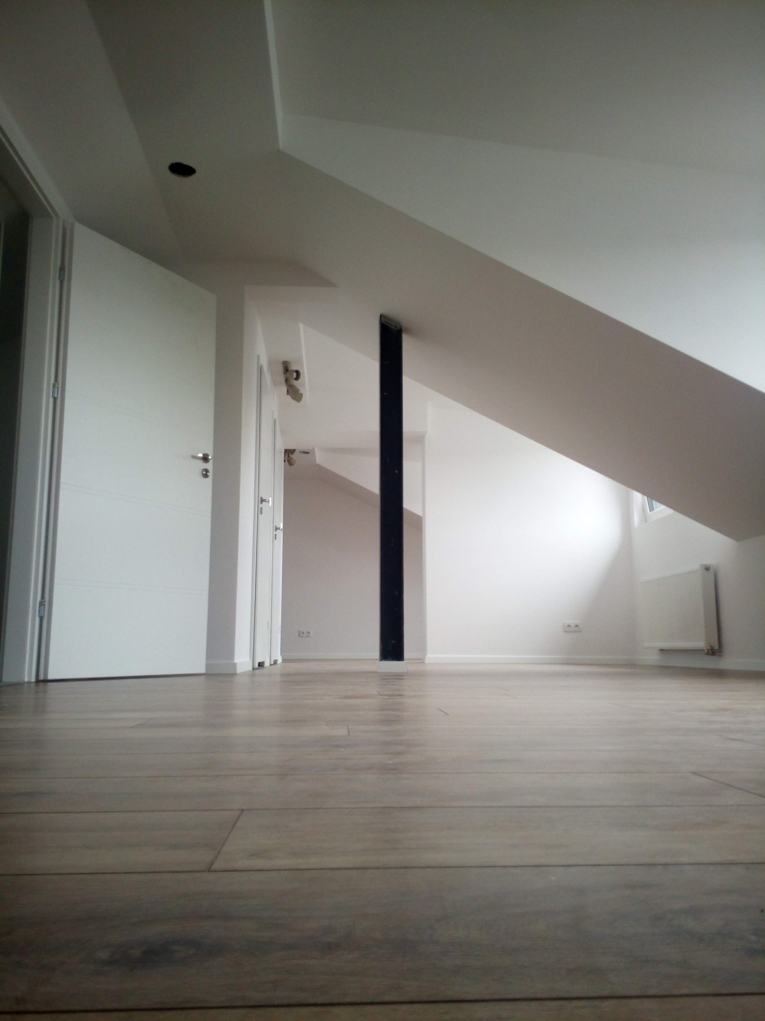 Poddasze po remoncie: świeżo ułożone panele podłogowe, białe ściany i drzwi, czarna kolumna, widok z poziomu podłogi.