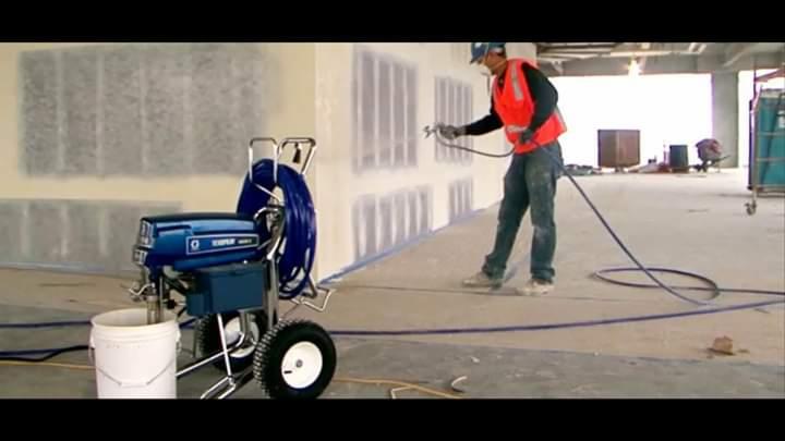 Pracownik w kasku i kamizelce odblaskowej maluje ścianę w surowym wnętrzu za pomocą profesjonalnego agregatu malarskiego, widoczne ślady szpachlowania i taśmy malarskiej.