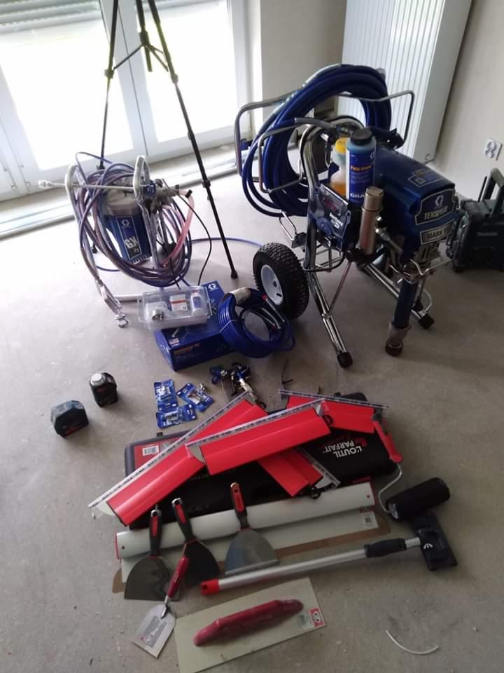 Profesjonalny sprzęt malarski: agregat hydrodynamiczny TexSpray Mark V, pistolet natryskowy na trójnogu, szpachle z czerwonymi uchwytami, listwy wykończeniowe i akcesoria na szarym podłożu w jasnym...