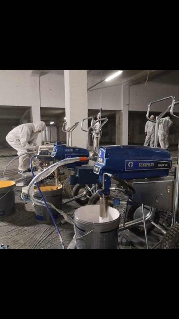 Pracownicy w kombinezonach przygotowują agregaty malarskie Texspray Mark VII z wiadrami farby w surowym wnętrzu, prawdopodobnie przed malowaniem natryskowym.