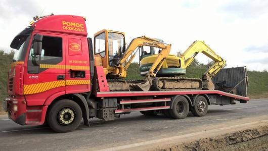 Czerwona laweta z napisem 'Pomoc Drogowa' przewozi dwie żółte, zakurzone mini-koparki Komatsu na niskopodwoziowej platformie.