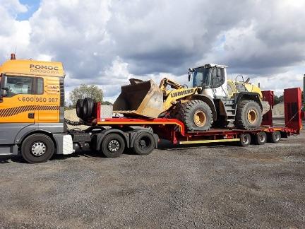 Żółto-szara ciężarówka pomocy drogowej z pomarańczową lawetą przewożącą żółto-białą ładowarkę Liebherr z brudną łyżką.