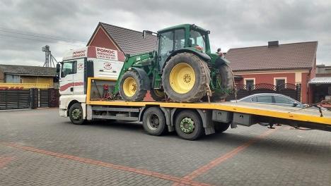 Ciężarówka z platformą przewozi zielony traktor John Deere na tle zabudowań.