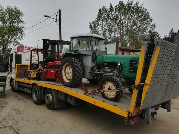 Żółta platforma ciężarówki przewozi stary, zielony traktor i czerwony wózek widłowy, zabezpieczone łańcuchami. W tle biała ciężarówka z napisem 'POMOC DROGOWA'.