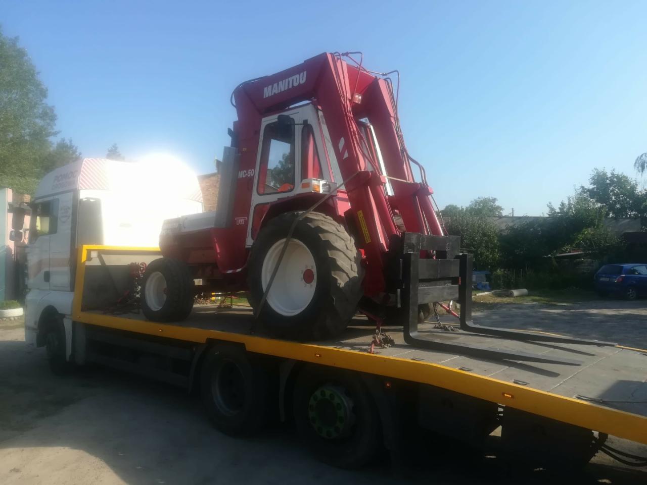 Czerwony wózek widłowy Manitou MC-50 z podniesionym masztem, zabezpieczony pasami na żółtej platformie ciężarówki pomocy drogowej z białym nadwoziem, na tle zieleni i błękitnego nieba.