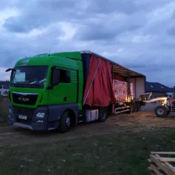 Zielona ciężarówka MAN z naczepą częściowo odsłoniętą, załadowana paletami dachówek, rozładowywana przez wózek widłowy na tle zabudowań mieszkalnych i pochmurnego nieba.