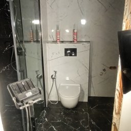 Grzegorz Omylski - Wnętrze łazienki w trakcie prac wykończeniowych, widoczna toaleta podwieszana, czarne płytki podłogowe z białym wzorem marmuru, białe płytki ścienne z szarym wzorem marmuru, reflektor budowlany...