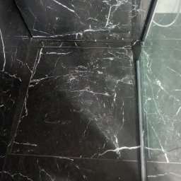 Grzegorz Omylski - Czarne marmurowe płytki podłogowe z białymi żyłkami w kabinie prysznicowej, widoczny odpływ liniowy i fragment buta.