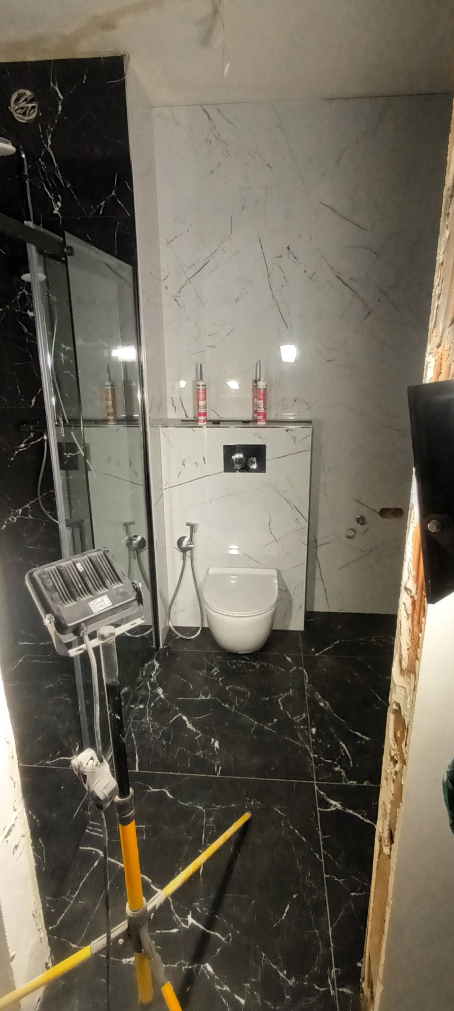 Wnętrze łazienki w trakcie prac wykończeniowych, widoczna toaleta podwieszana, czarne płytki podłogowe z białym wzorem marmuru, białe płytki ścienne z szarym wzorem marmuru, reflektor budowlany...