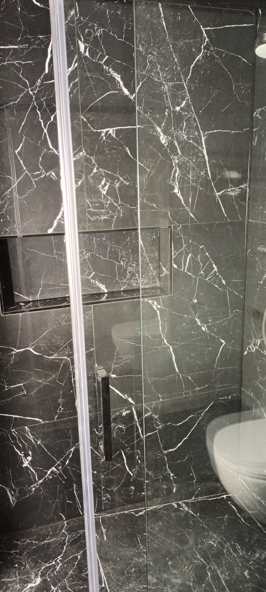 Wykończona łazienka z czarnymi płytkami imitującymi marmur, przeszklona kabina prysznicowa z czarnym uchwytem, wnęka w ścianie wyłożona tą samą płytką.