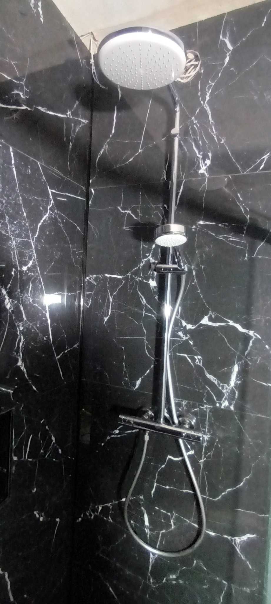 Prysznic z deszczownicą zamontowany na ścianie wyłożonej czarnymi płytkami imitującymi marmur z białymi żyłami.