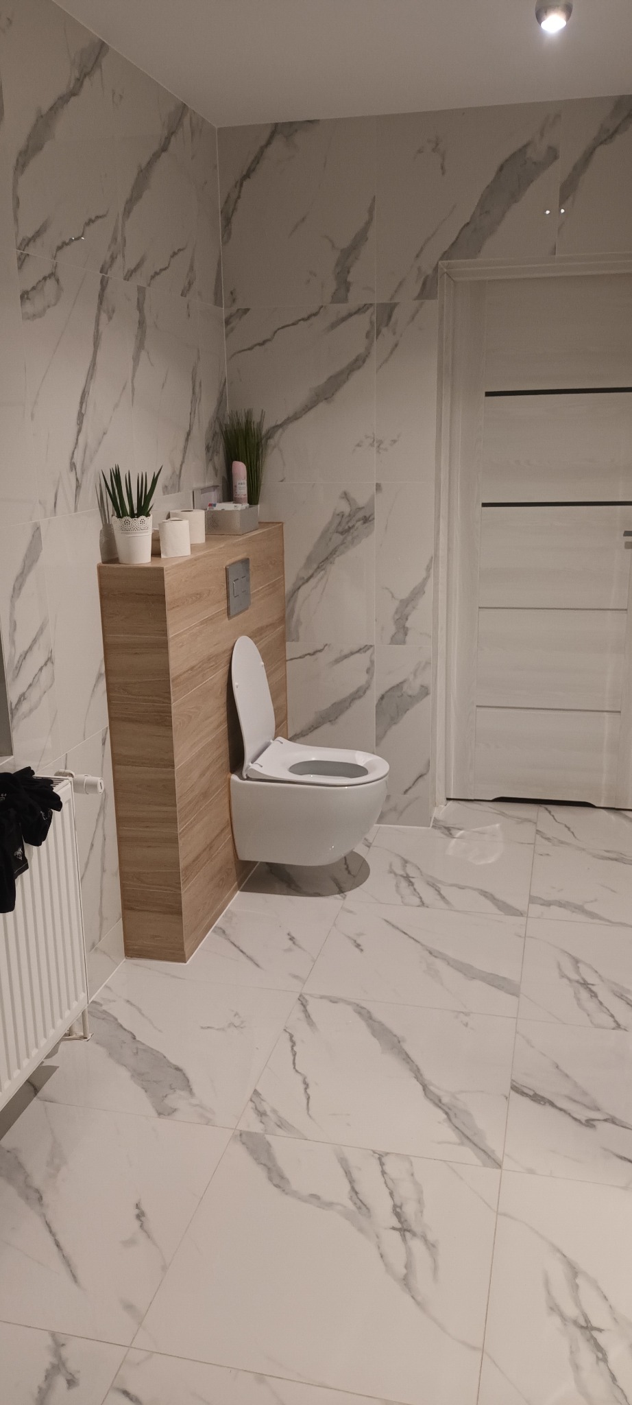 Nowoczesna łazienka z podwieszaną toaletą, marmurowe płytki na ścianach i podłodze, drewniana zabudowa stelaża WC z półką na akcesoria, białe drzwi w tle.