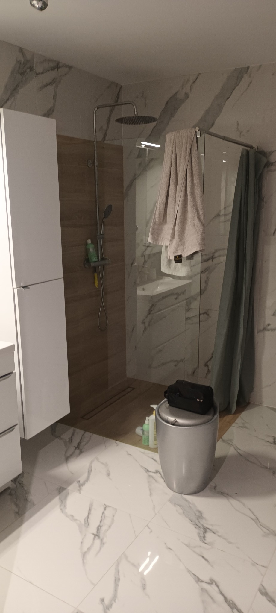 Nowoczesna łazienka z kabiną prysznicową typu walk-in, marmurowymi płytkami na ścianach i podłodze, białymi szafkami, deszczownicą i ręcznikami.