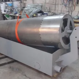 Metalowy, cylindryczny bęben przemysłowy z silnikiem, zabezpieczony pomarańczowym pasem transportowym, umieszczony na szarej, metalowej podstawie, w warsztatowym otoczeniu.