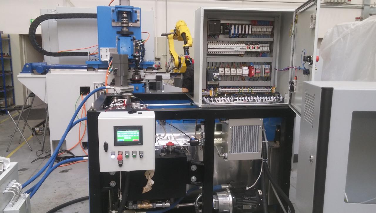 Zautomatyzowana linia produkcyjna z robotem przemysłowym i szafą sterowniczą z otwartymi drzwiami, widoczne elementy hydrauliczne i panel sterowania z zielonym ekranem.