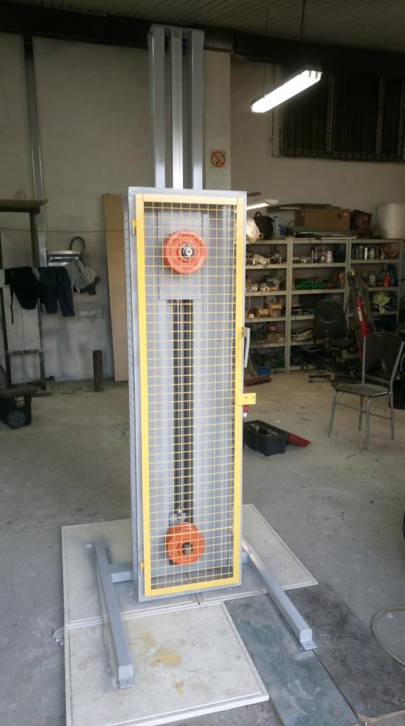 Test stanowiskowy do bram garażowych: metalowa konstrukcja z żółtymi elementami zabezpieczającymi, widoczne obciążniki i linki, szara podstawa na betonowej posadzce w warsztacie.