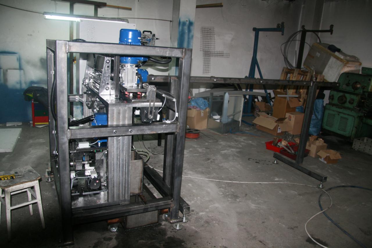 Surowa konstrukcja automatyki do bram w warsztacie, widoczne silniki, łańcuchy i metalowa rama na kółkach, z narzędziami na stołku w tle.