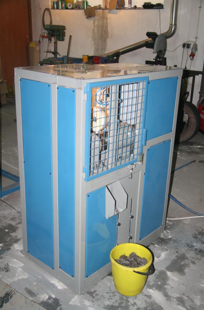 Niebiesko-szara obudowa maszyny z otwartą kratą, przez którą widać wewnętrzne elementy, z żółtym wiadrem pełnym metalowych elementów u dołu.