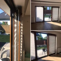 Widok z bliska na mechanizm przesuwny i zamknięte poziome pasy materiału w oknie tarasowym, z widokiem na ogr&oacute;d i dom w tle, ujęcie z wnętrza pomieszczenia.