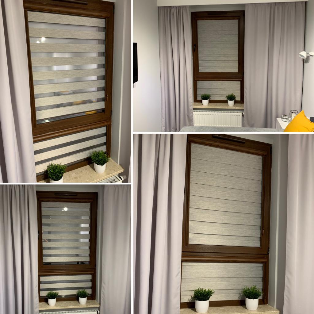 Okna z brązowymi ramami i szaro-białymi przesłonami okiennymi w pokoju z szarymi zasłonami i dekoracyjnymi roślinami w doniczkach na parapecie.