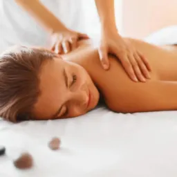 Kobieta relaksuje się podczas masażu pleców, z kamieniami ułożonymi na prześcieradle w tle.